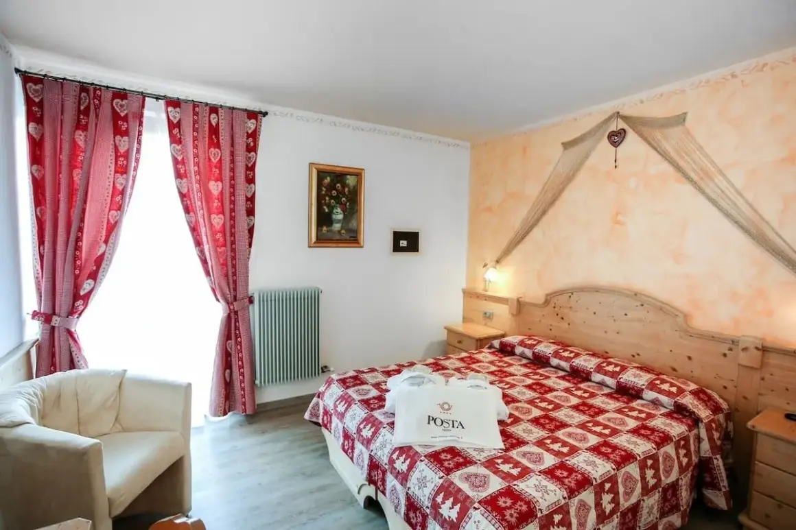 Camera accogliente con letto a quadri rossi, tende a cuoricini e un sacchetto con logo POSTA HOTEL 1899.