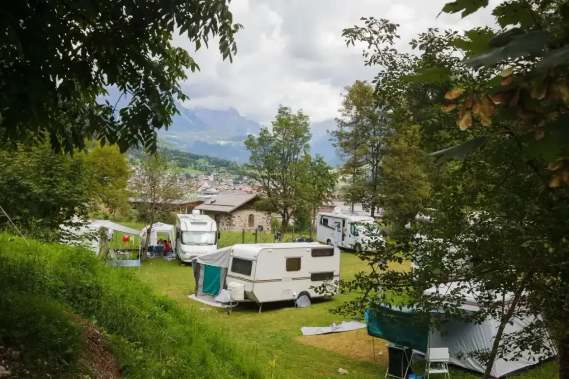Un tranquillo camping calavise con roulotte e camper immersi nel verde, sullo sfondo un villaggio alpino e montagne nebbiose.