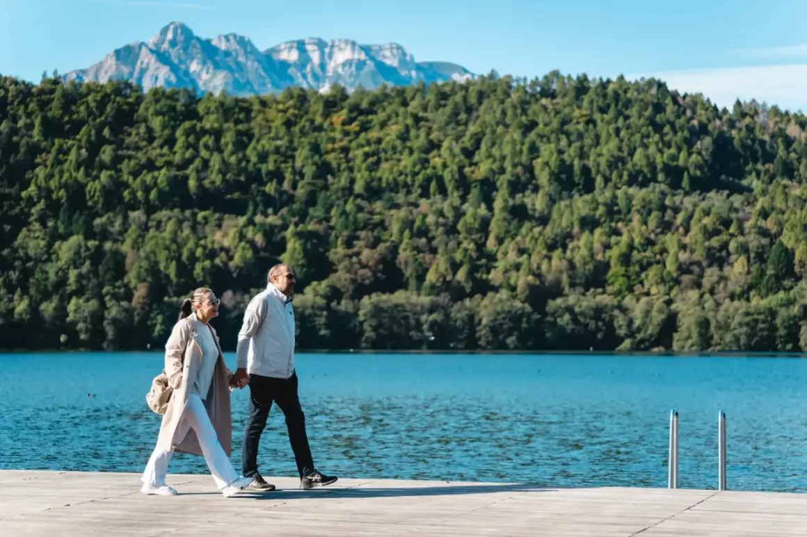 Coppia passeggia mano nella mano sul sentiero dei pescatori a Levico, con lago e montagne sullo sfondo.