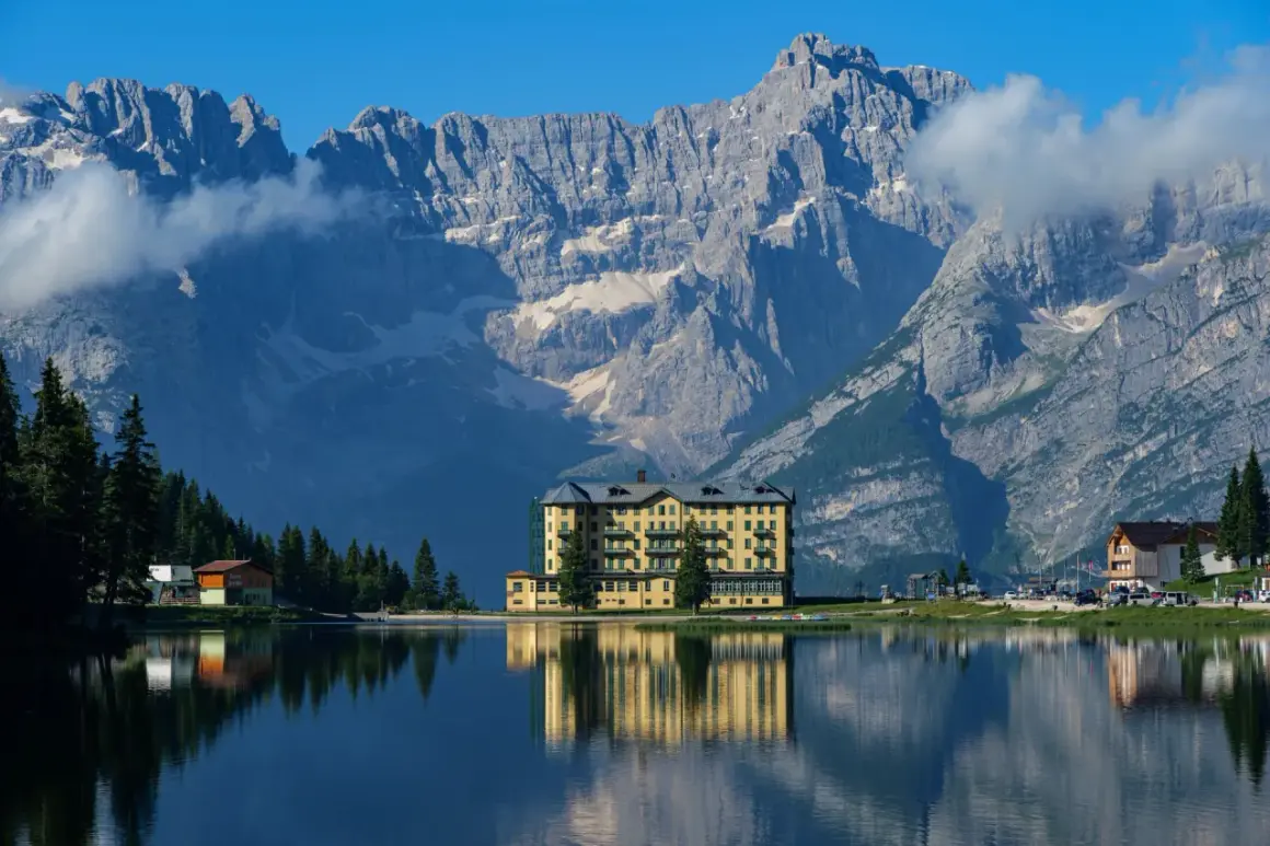 Un hotel giallo si specchia in un lago alpino, con imponenti montagne sullo sfondo: ecco dove si trovano le Dolomiti.