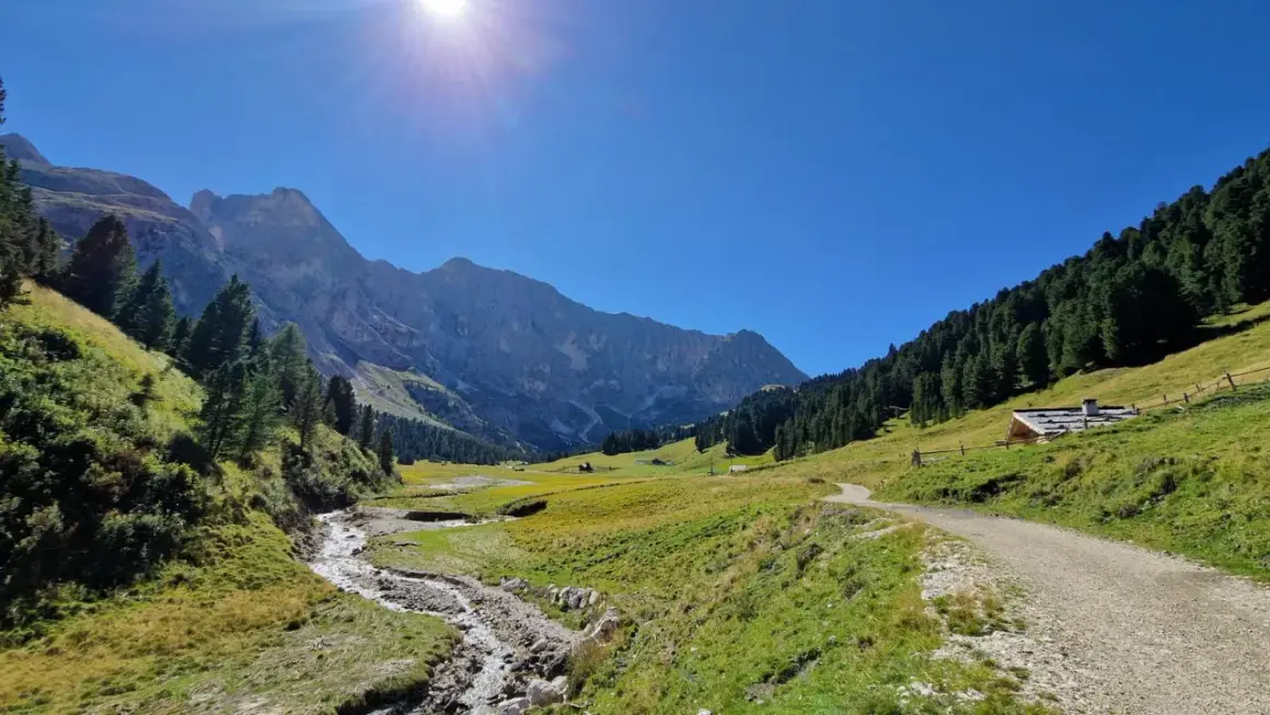 Sentiero sterrato che sale verso un rifugio alpino, con un ruscello che scorre in primo piano e montagne imponenti sullo sfondo, tipico del passo Duron.