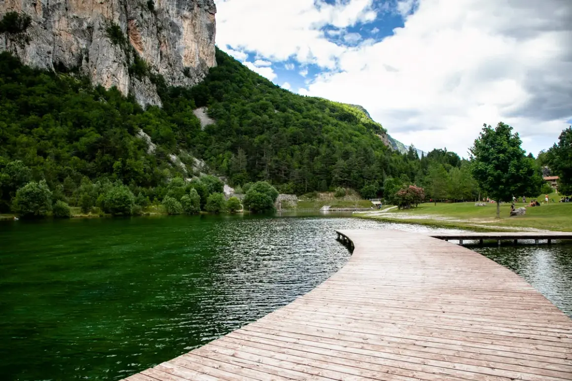 Un pontile in legno curva dolcemente sul lago di Nembia, circondato da montagne verdi e un cielo nuvoloso.