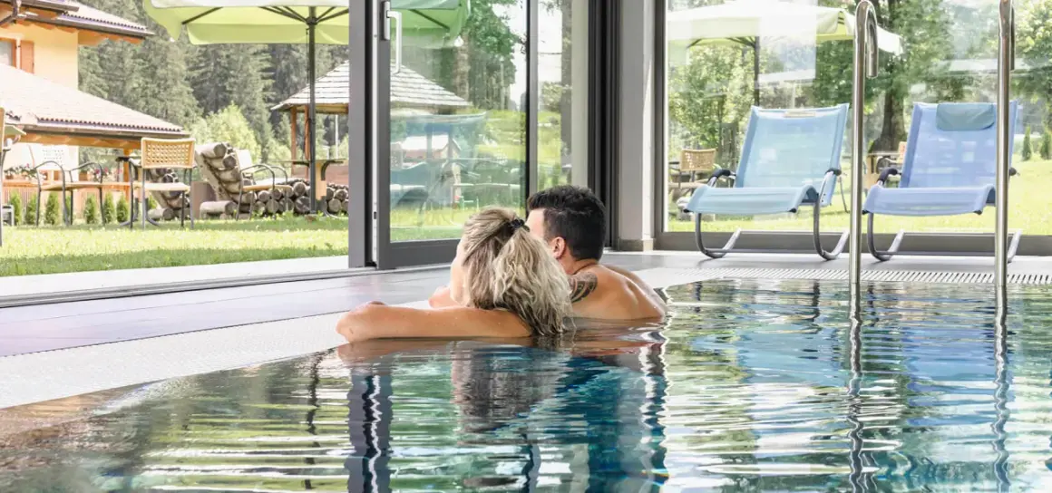 Coppia rilassata nella piscina interna di un wellness hotel Dolomia, con vista sul giardino e sulle montagne.