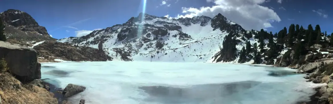 Il lago delle stellune, ghiacciato e circondato da montagne innevate, riflette il cielo azzurro e le nuvole.