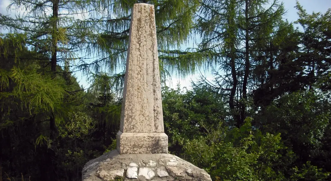 Obelisco in pietra sul **monte Zugna**, circondato da alberi di larice e vegetazione rigogliosa.