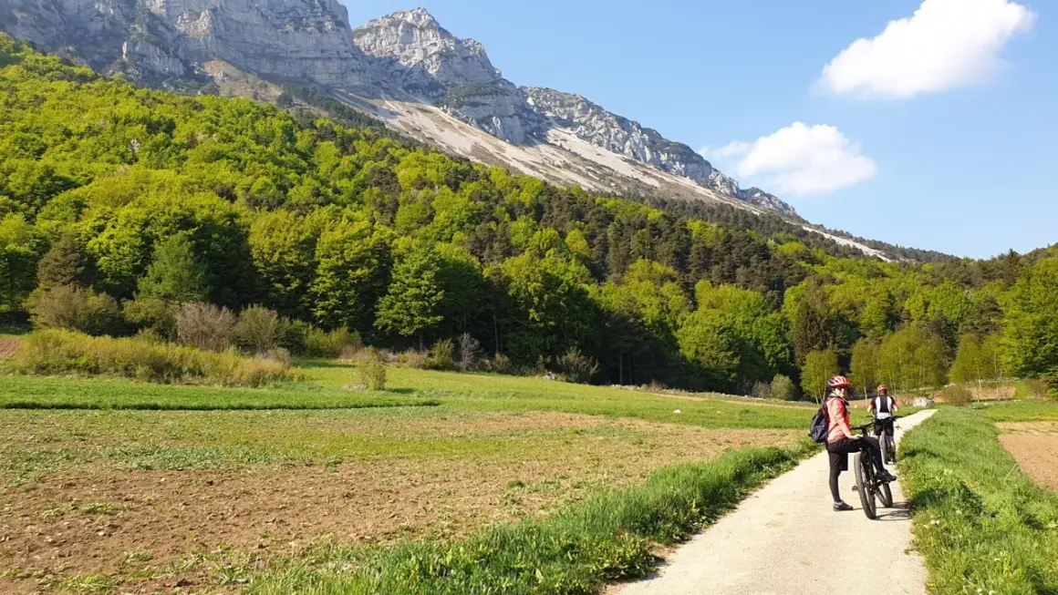 Ciclisti su un sentiero sterrato, con campi coltivati e un bosco rigoglioso ai piedi di una montagna imponente. Un passo bordala da godersi in bicicletta.