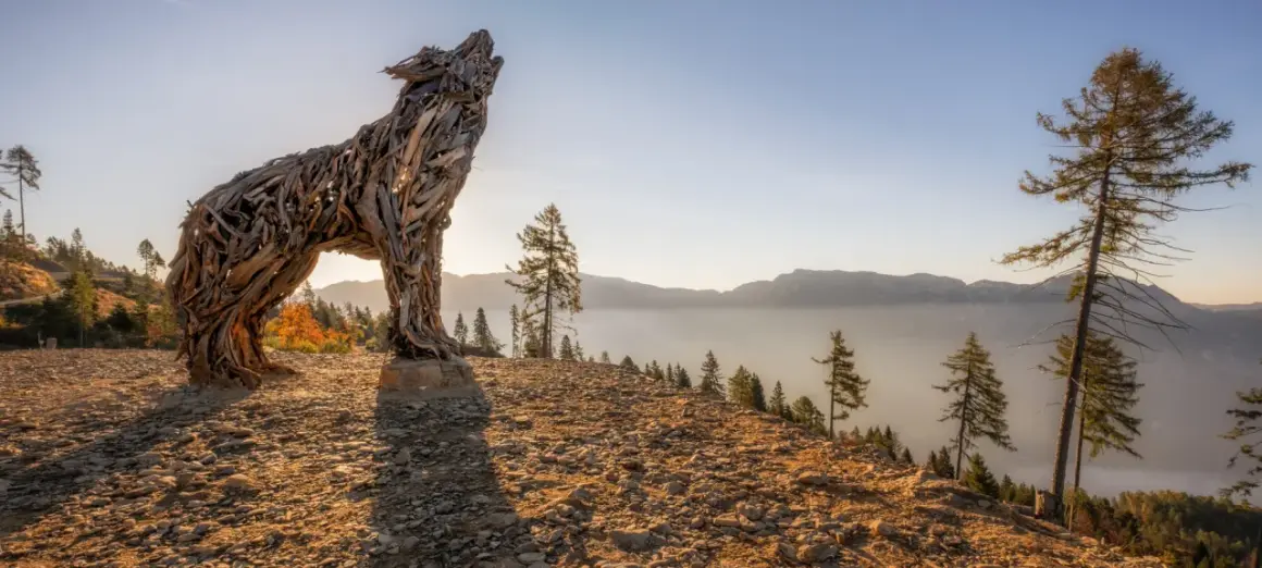Scultura di una lupa del Lagorai in legno, che ulula al tramonto su un crinale roccioso, con montagne nebbiose all'orizzonte.