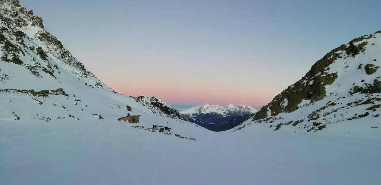 Bivacco Paolo e Nicola, un piccolo rifugio solitario tra montagne innevate al tramonto, con un cielo rosa e azzurro.