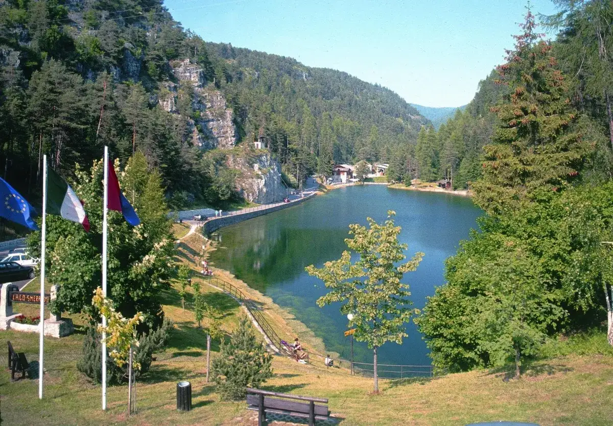 Il lago smeraldo incastonato tra montagne boscose, con case e un muretto che lo costeggiano. Bandiere sventolano in primo piano.