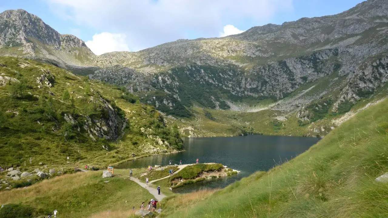 Un sentiero serpeggia tra prati verdi e rocce, conducendo a un lago alpino. Persone passeggiano, godendosi il panorama durante il giro dei 5 laghi.