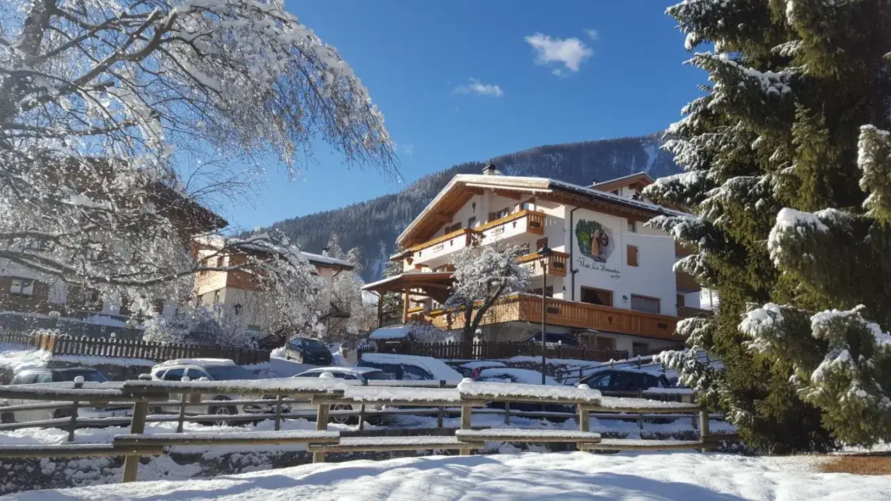 Hotel La Romantica Moena, immerso nella neve, con alberi innevati e un cielo azzurro brillante. Un'atmosfera da fiaba invernale.