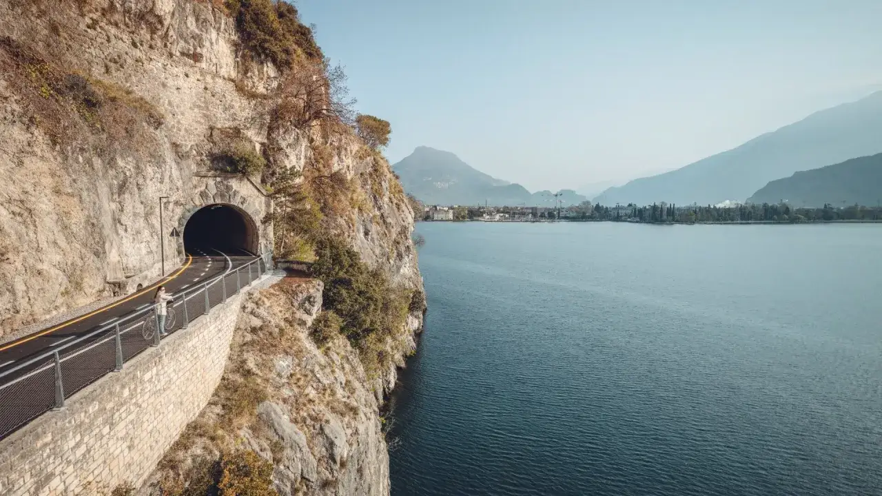 Ciclabili Trentino: una ciclista ammira il lago da una strada scavata nella roccia, con un tunnel in primo piano.