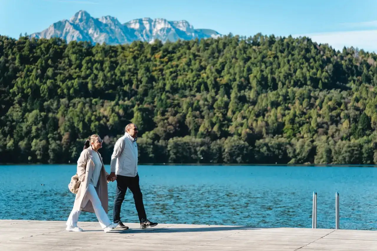 Coppia passeggia mano nella mano sul sentiero dei pescatori a Levico, con il lago e le montagne sullo sfondo.