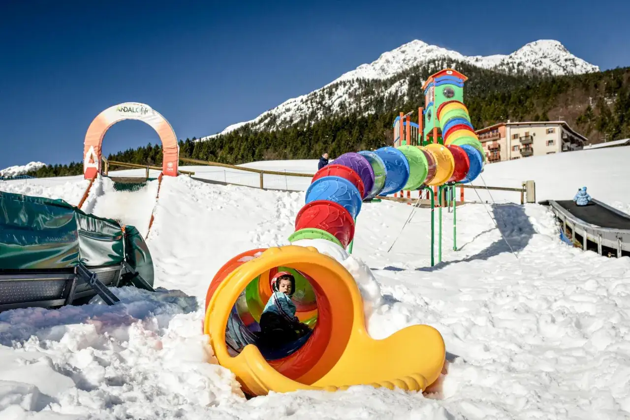 Bambino gioca nel colorato scivolo del Pejo Kinderland innevato, con montagne sullo sfondo.