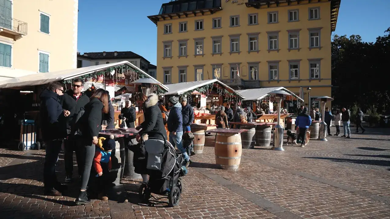 Gente al markt Riva del Garda, tra bancarelle addobbate e botti di legno, sotto un cielo azzurro.