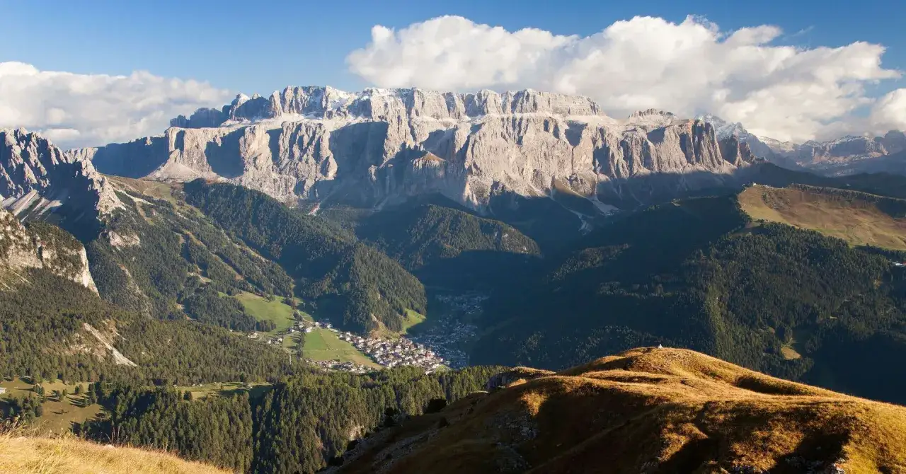 Villaggio alpino incastonato tra boschi e montagne imponenti, con il passo Sella sullo sfondo.