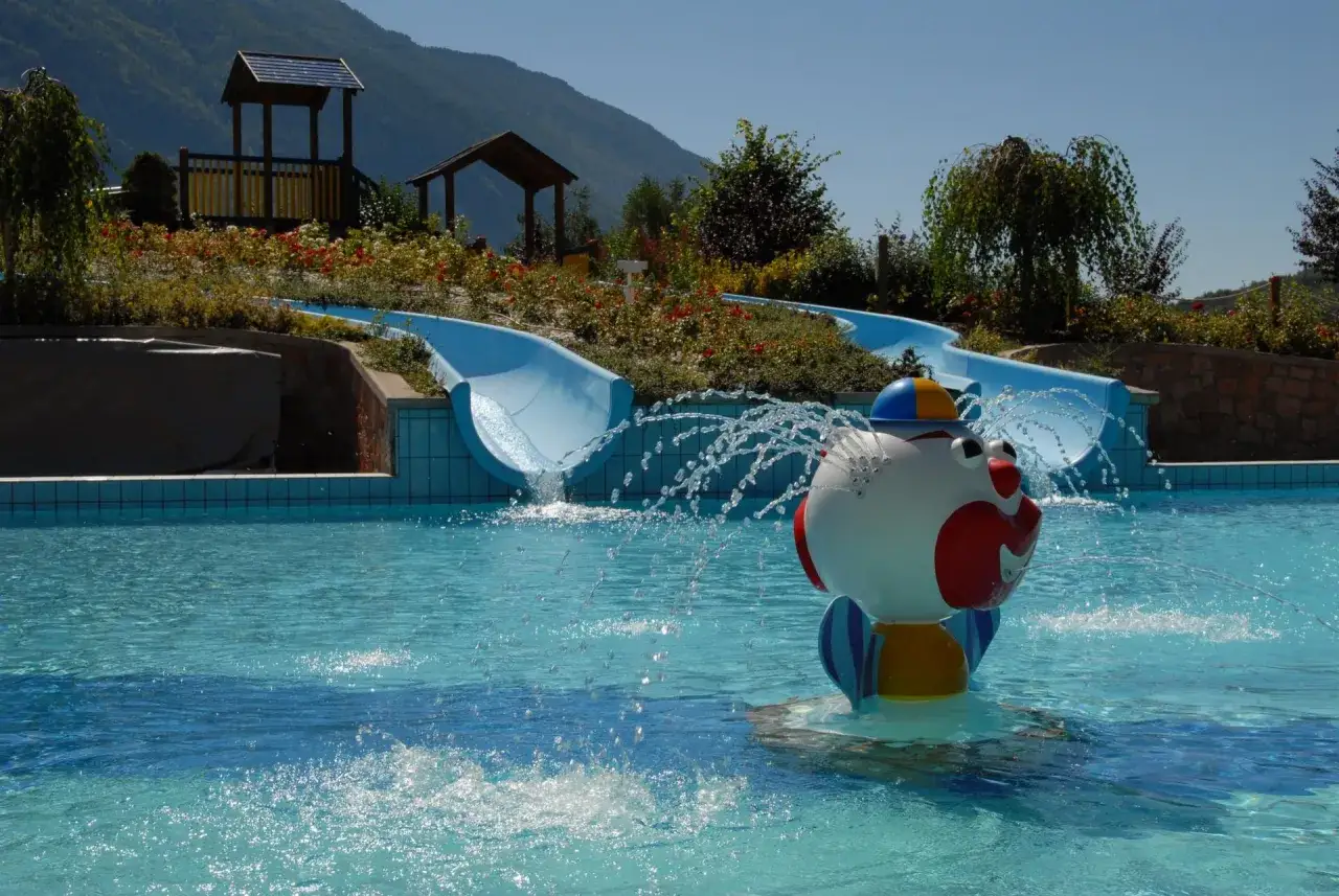 Acquapark Molveno: scivoli azzurri e un simpatico spruzzatore a forma di clown in una piscina cristallina, con montagne sullo sfondo.