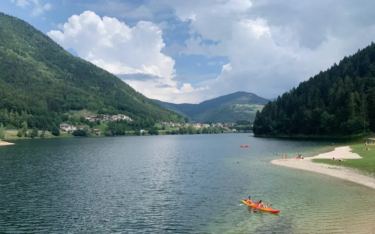 Un kayak giallo e rosso solca le acque limpide del lago di Pinè, circondato da montagne verdi e un villaggio pittoresco.