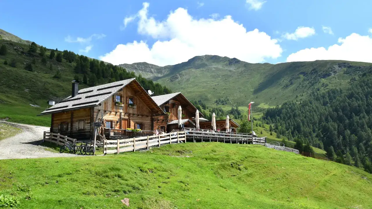 Malga Campo, un rifugio alpino in legno con terrazza e ombrelloni, immerso nel verde dei prati e circondato da montagne boscose.