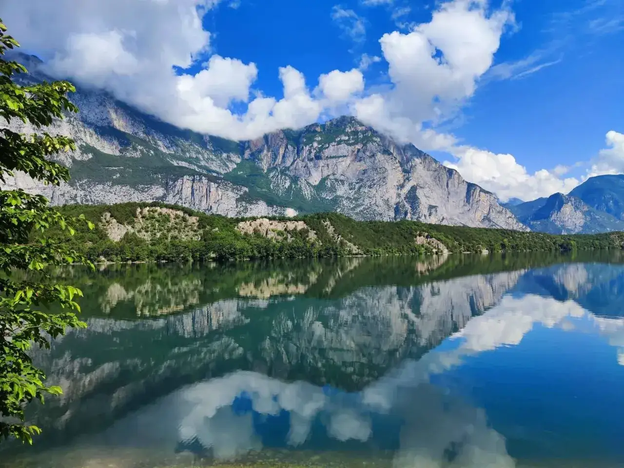 Il lago di Cavedine riflette montagne imponenti e nuvole bianche in un cielo azzurro.
