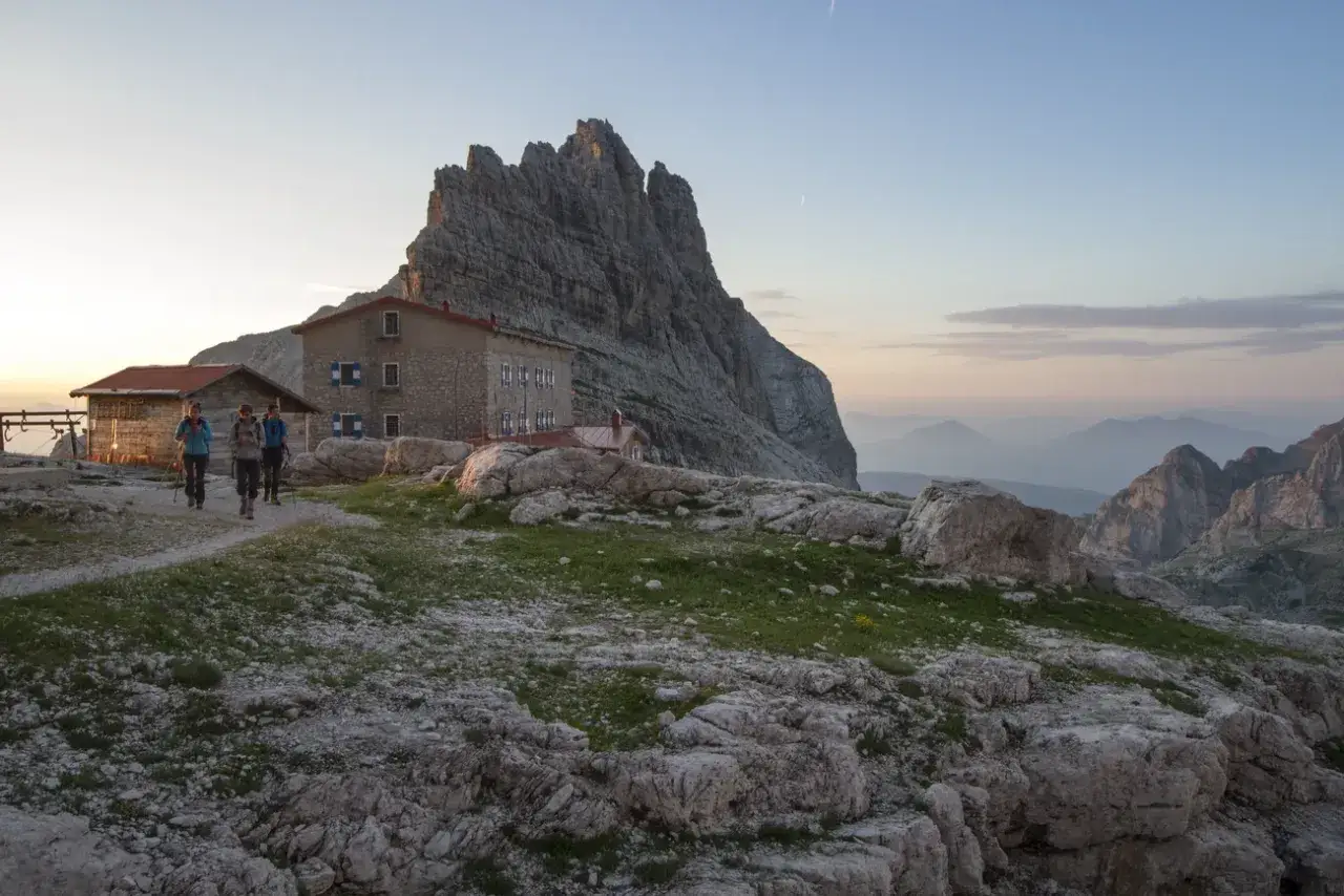 Alba sulle ferrate Dolomiti: escursionisti si avvicinano a un rifugio alpino con imponente parete rocciosa sullo sfondo.