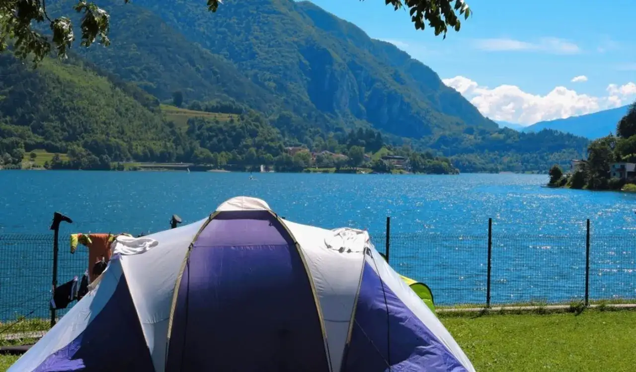 Tenda viola e bianca su prato verde, lago azzurro e montagne sullo sfondo. Il best Ledro camping per una vacanza indimenticabile.
