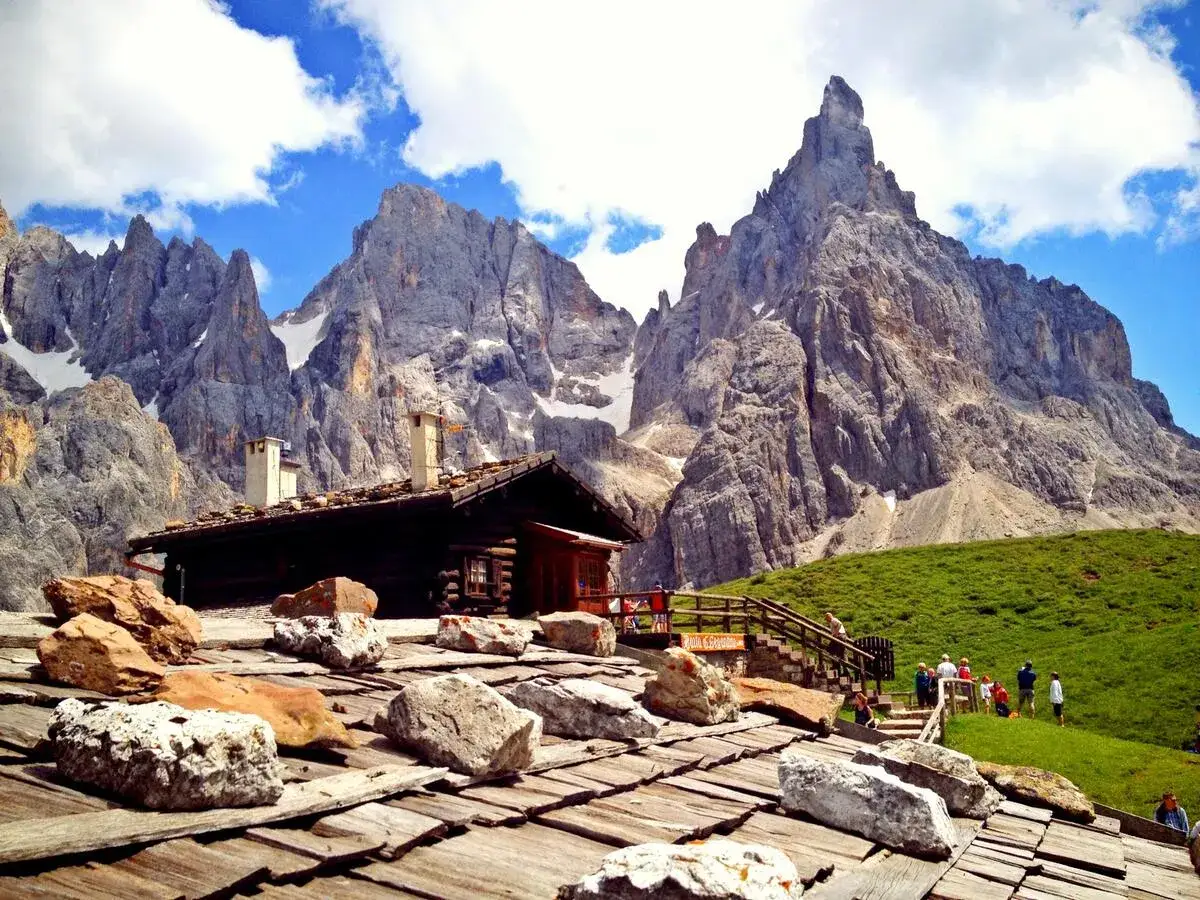 La baita Segantini, un rifugio alpino in legno, si erge maestosa ai piedi di imponenti vette rocciose. Un prato verde e un cielo azzurro completano il paesaggio.