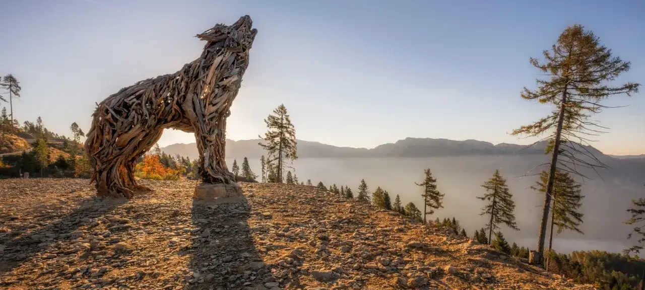Scultura di una lupa del Lagorai in legno, che ulula al sole del mattino su un crinale roccioso con vista su montagne nebbiose.