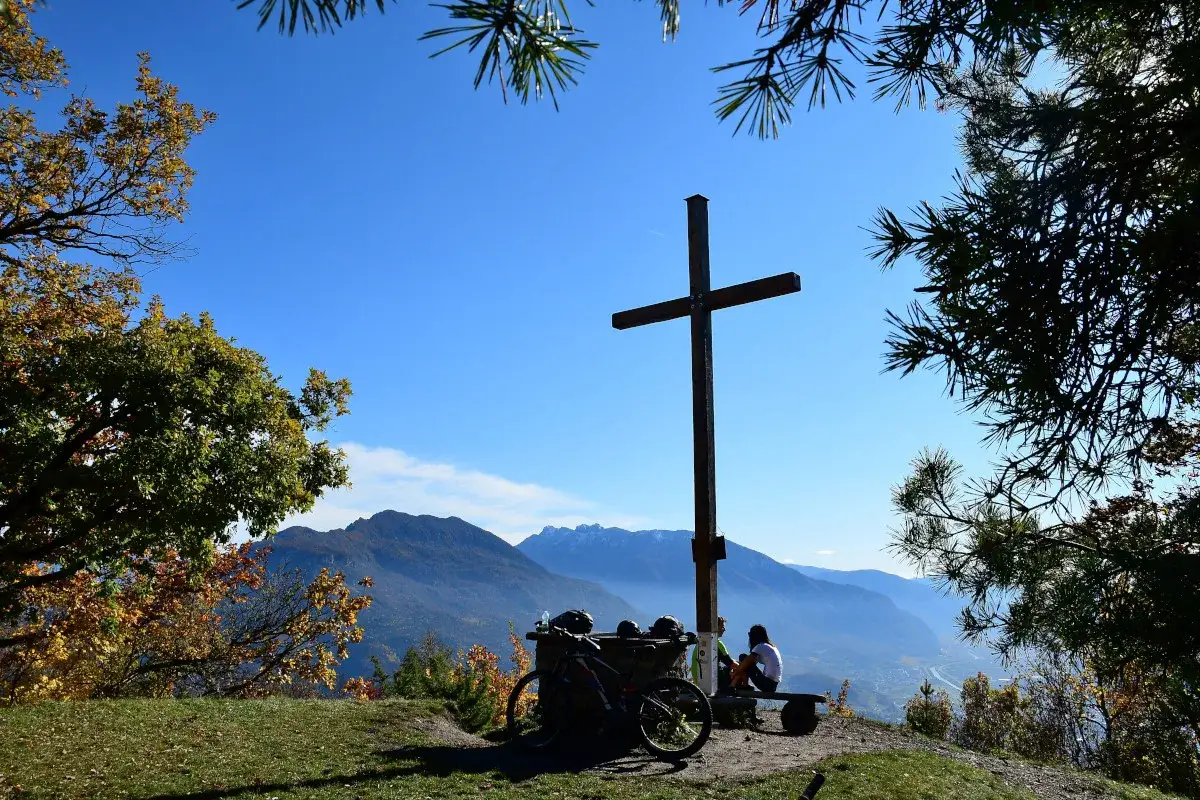 Croce sul monte Calisio, con ciclista che riposa e vista panoramica sulle montagne.