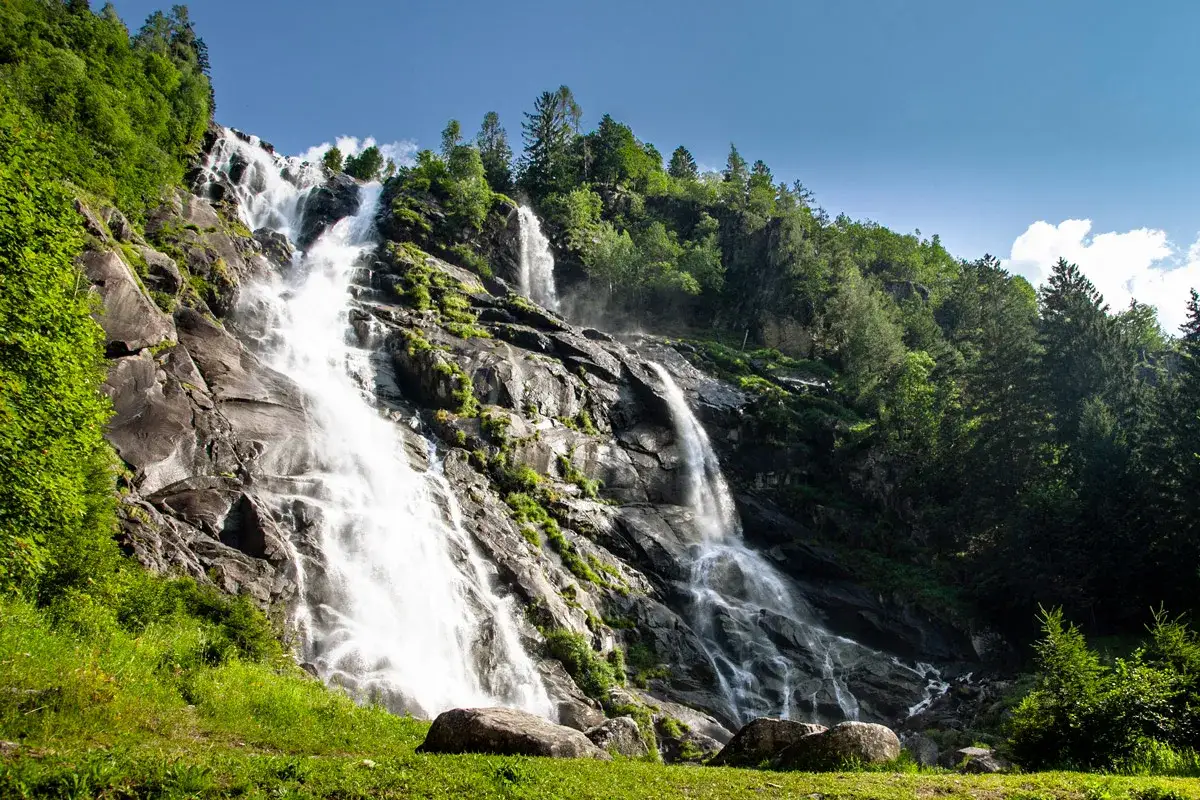Cascate maestose nel Val di Genova, con acqua che scende impetuosa su rocce scure, circondate da fitti boschi verdi e un cielo azzurro.
