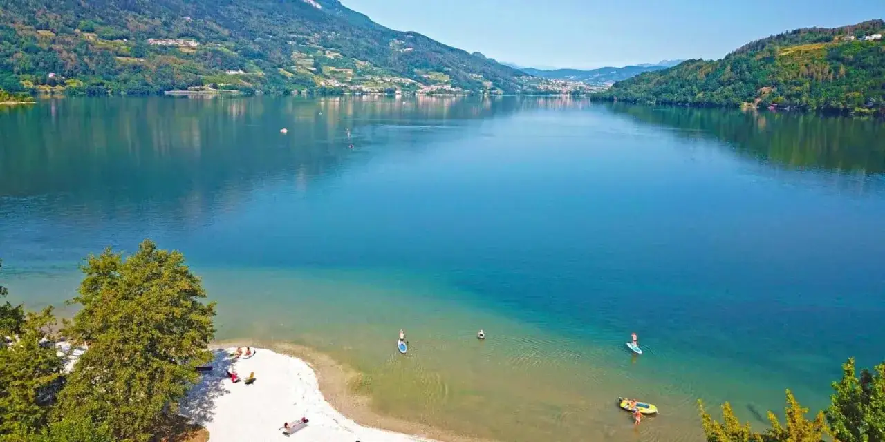 Spiaggia e acque cristalline del lago di Caldonazzo, con persone che praticano paddleboard e si rilassano sulla riva.