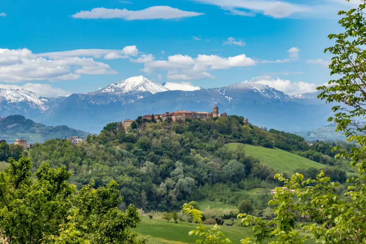 Un borgo arroccato su una collina, con montagne innevate sullo sfondo. Un paesaggio da sogno per un viaggio nelle Marche.