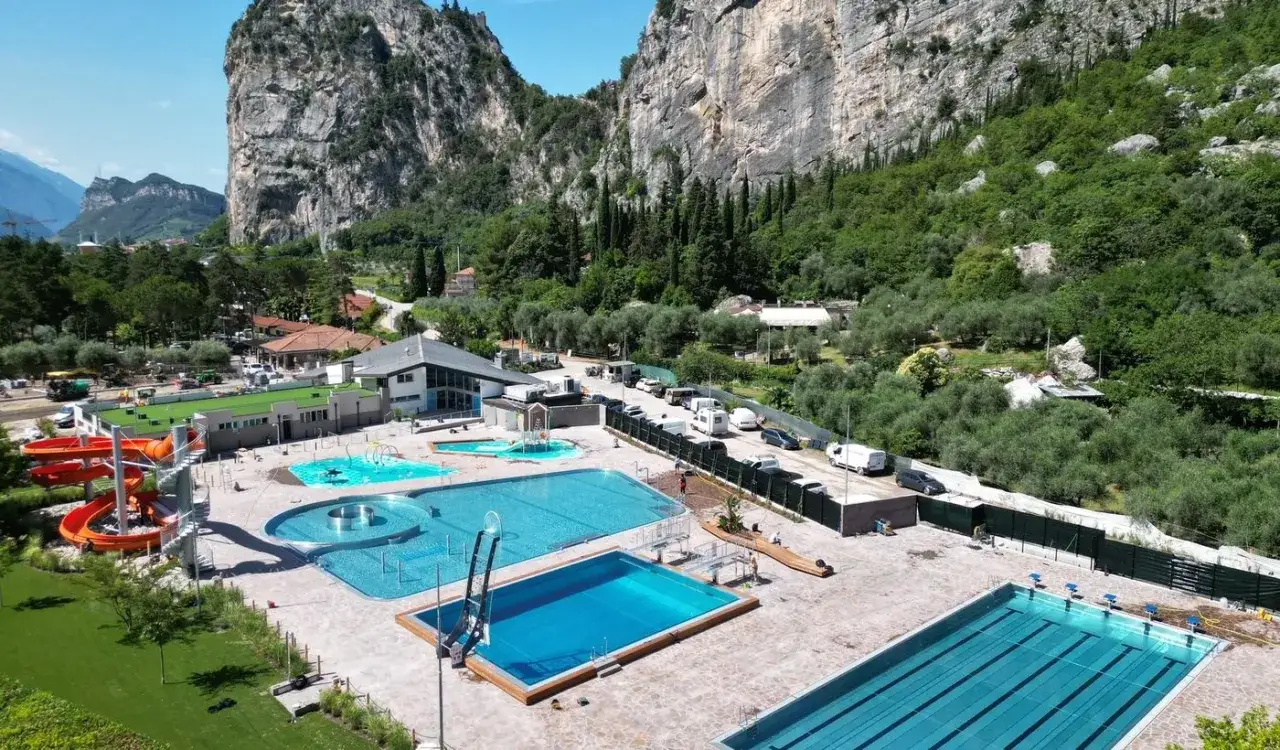 Un complesso acquatico moderno con scivoli arancioni, piscine di varie forme e un campo da nuoto, incastonato tra montagne verdi e uliveti, ideale per un soggiorno in camping ad Arco.