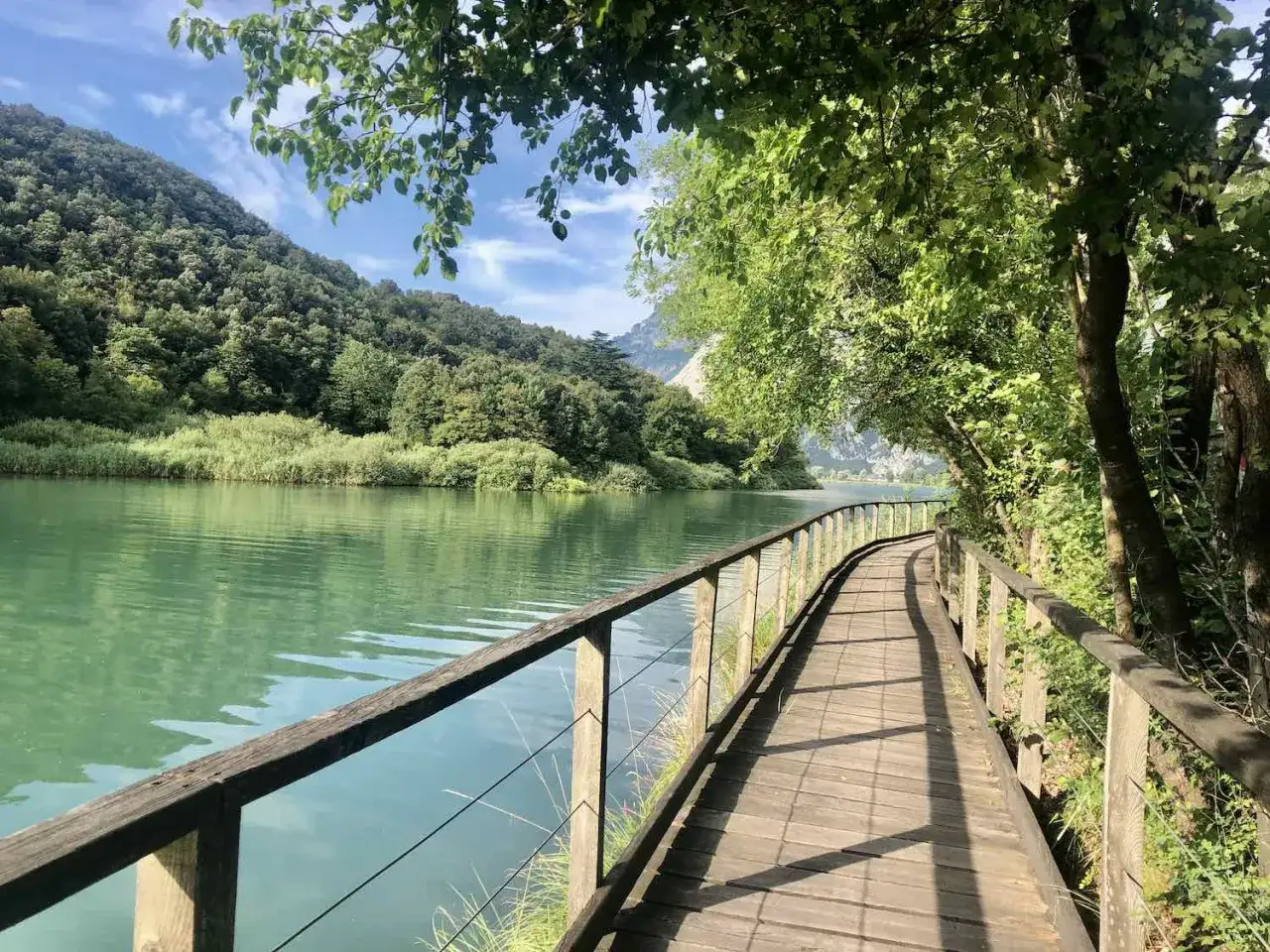 Sentiero in legno lungo il lago di Toblino, circondato da vegetazione lussureggiante e montagne verdi.