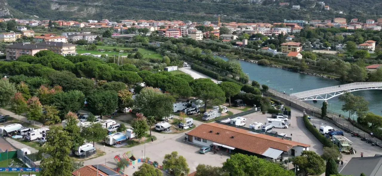 Campeggio con roulotte e camper vicino al fiume, con un ponte bianco e la città di Arco sullo sfondo.