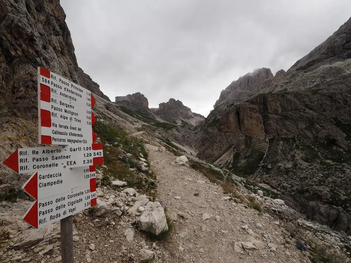 Cartello con indicazioni per il Passo Principe e altre destinazioni in montagna. Sentiero roccioso sale tra le vette.
