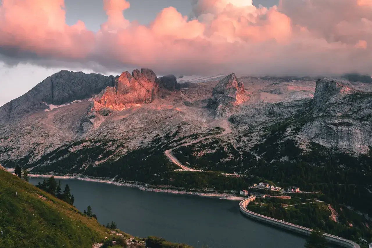 Alba rosata sulle Dolomiti al Passo Fedaia, con il lago artificiale e la diga che riflettono il cielo infuocato.
