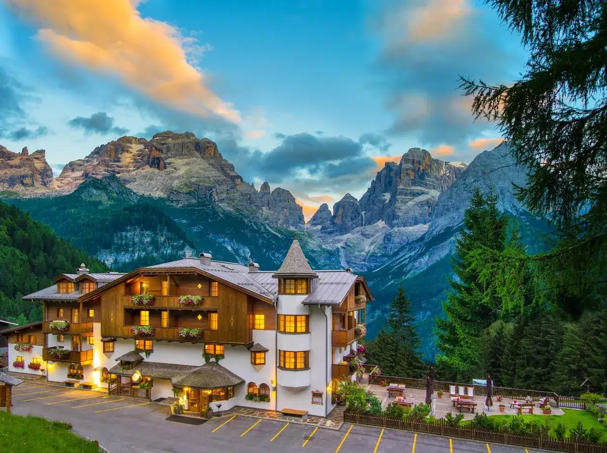 L'albergo Miramonti, con le sue finestre illuminate, si staglia maestoso ai piedi di imponenti vette montuose sotto un cielo al tramonto.