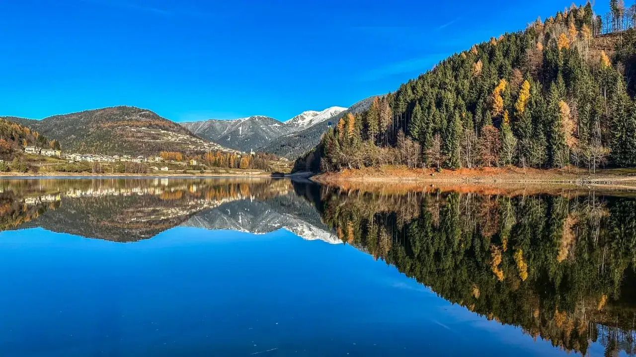 Il lago delle Piazze riflette montagne innevate e boschi autunnali sotto un cielo azzurro.