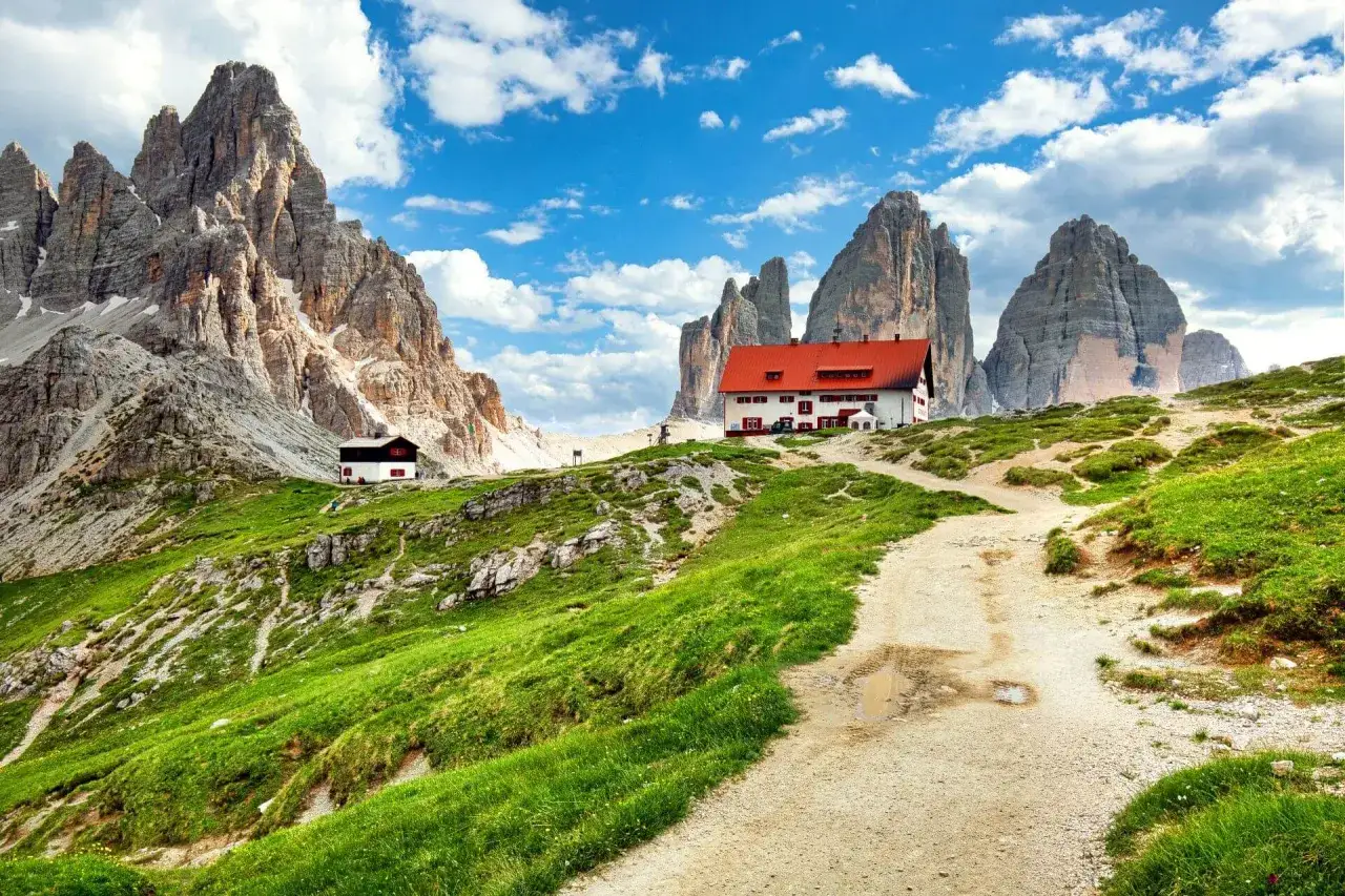 Un rifugio con tetto rosso domina un prato verde ai piedi di imponenti vette rocciose. Perfetto per un weekend in montagna.
