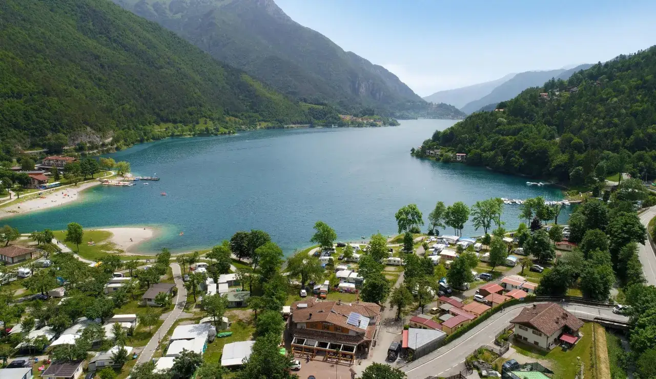 Vista aerea del camping Riviera Caldonazzo, con case mobili, roulotte e bungalow affacciati sul lago azzurro e circondati da montagne verdi.