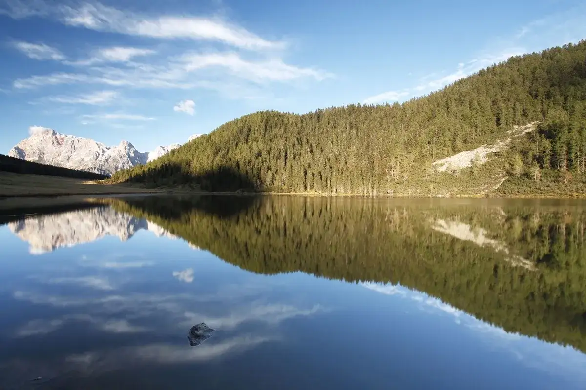Riflessi perfetti nel lago di Calaita: montagne innevate e boschi di conifere si specchiano nell'acqua calma sotto un cielo azzurro con nuvole bianche.
