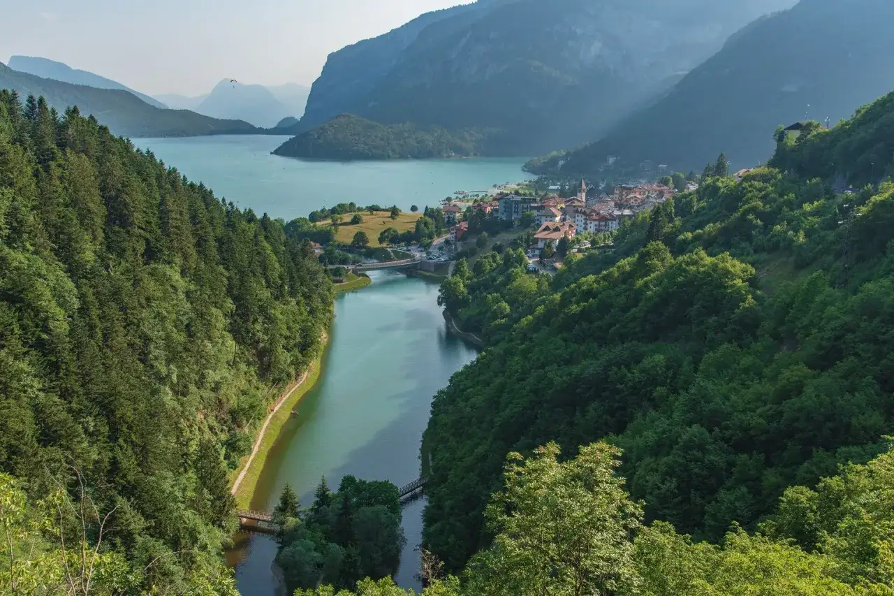 Panoramica mozzafiato del lago di Molveno, incastonato tra montagne verdi e un borgo pittoresco. Un parapendio vola nel cielo azzurro.
