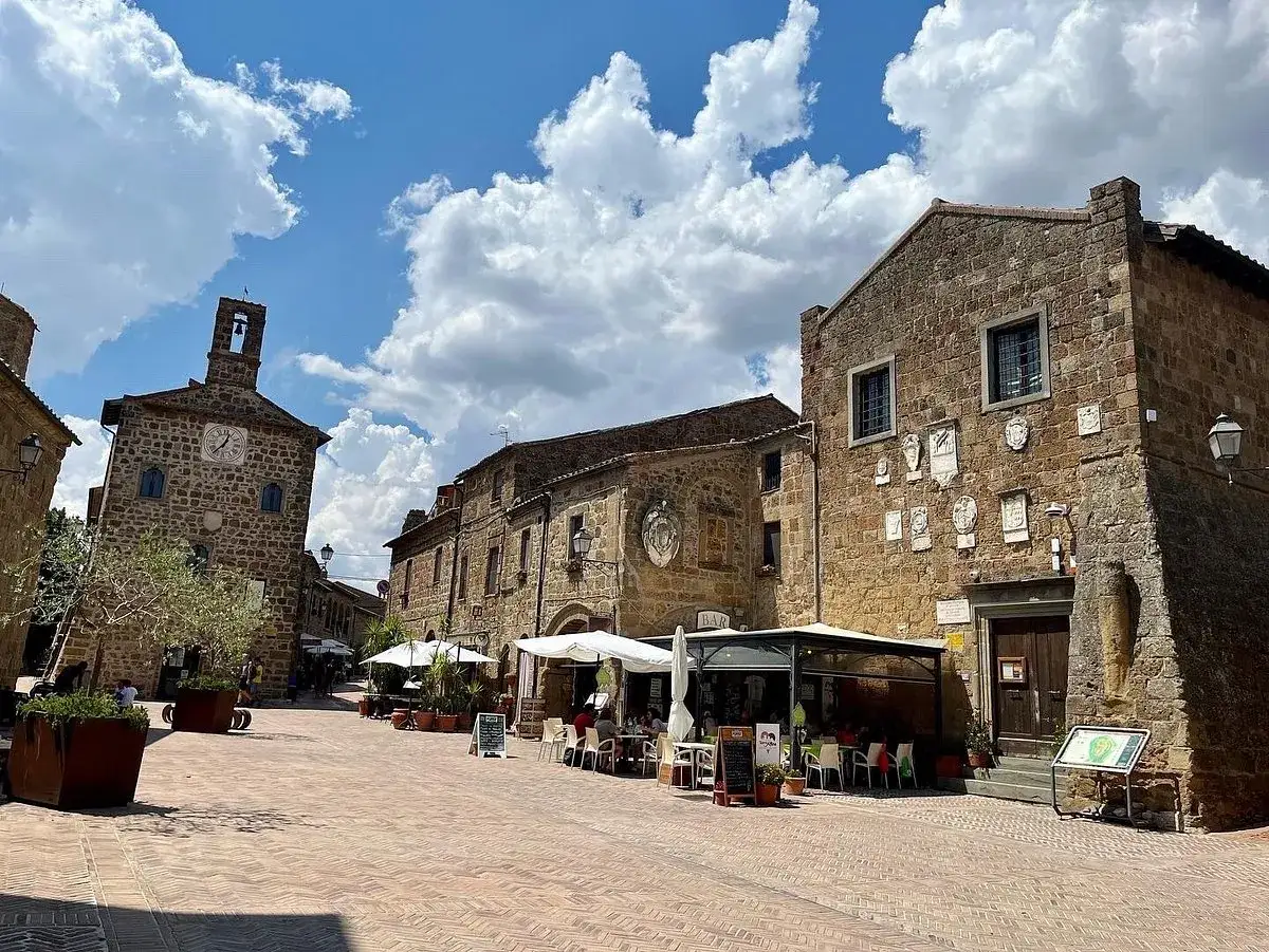 Piazza medievale con edifici in pietra, un campanile con orologio e tavolini all'aperto. Un luogo incantevole dove scansano cosa vedere.