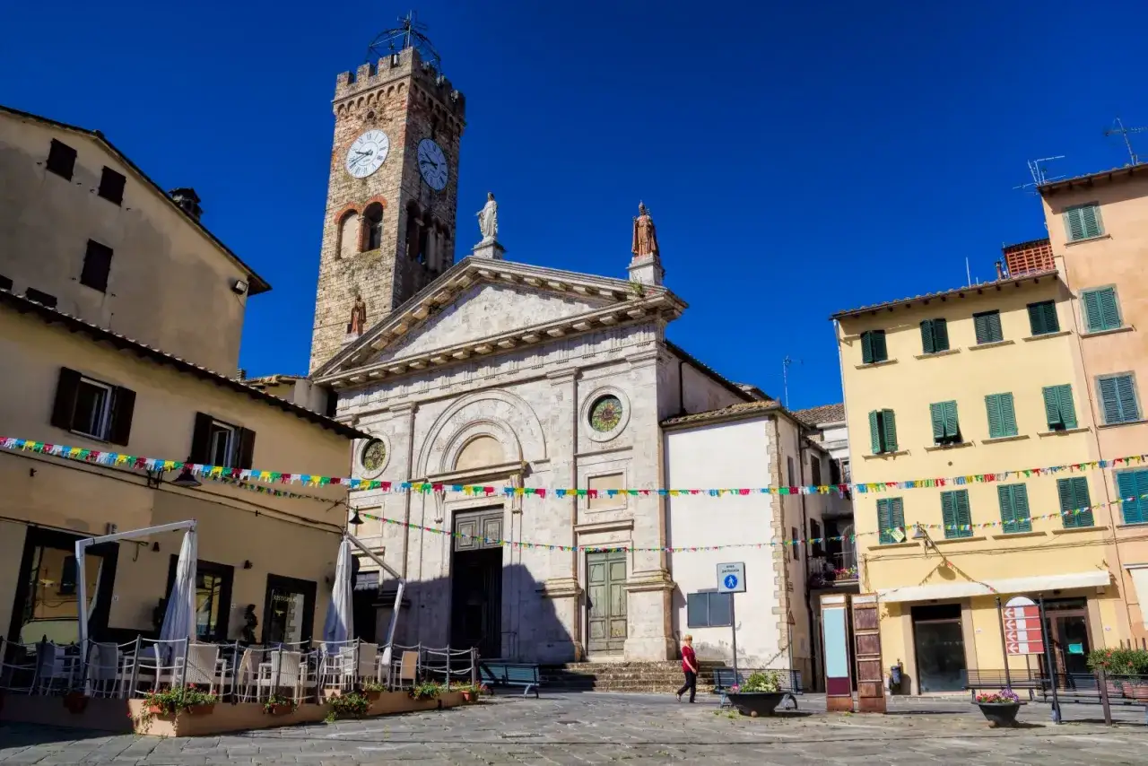 Piazza a Poggibonsi cosa vedere: chiesa con torre campanaria, case colorate e tavolini all'aperto.