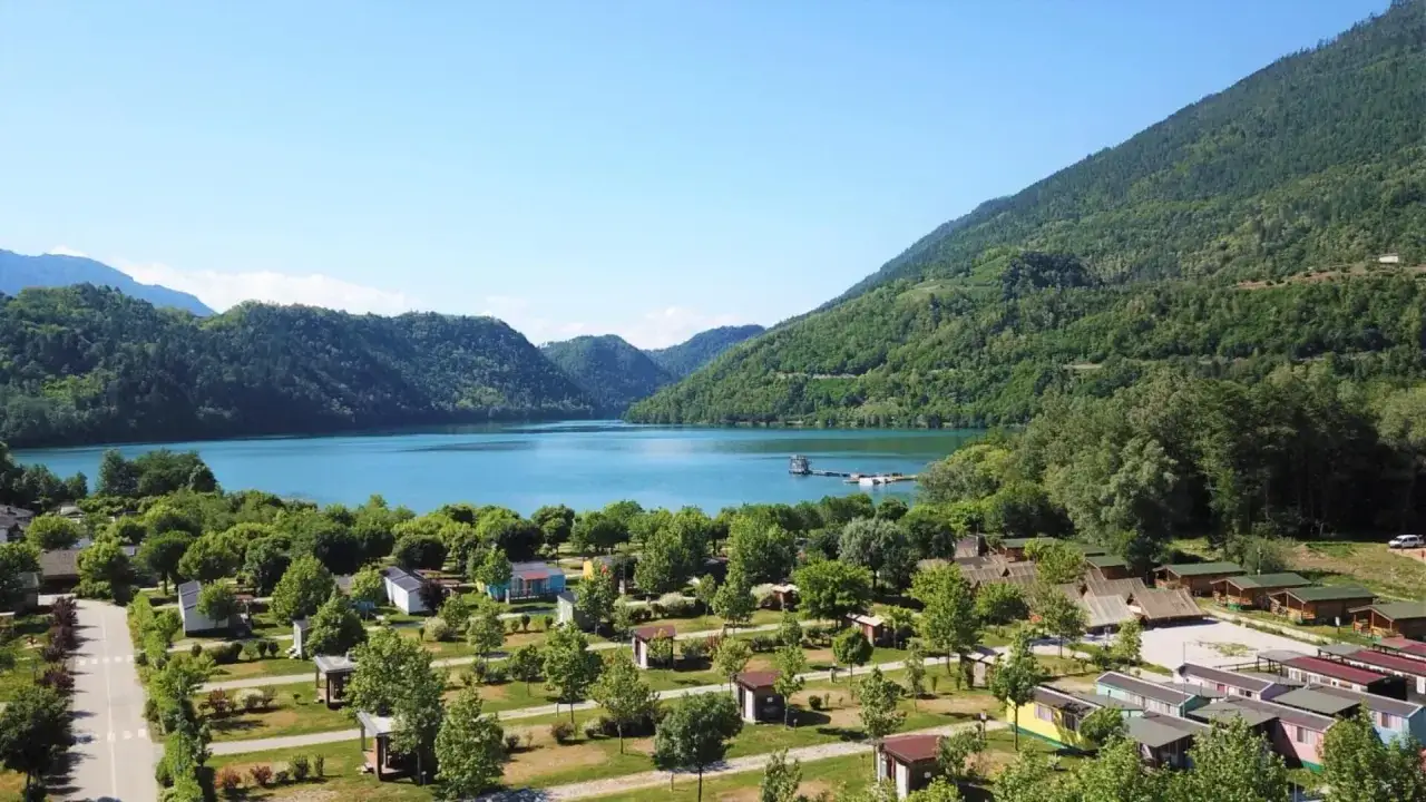 Un incantevole lago Levico camping village immerso nel verde, con bungalow colorati e il lago azzurro che si estende tra le montagne.