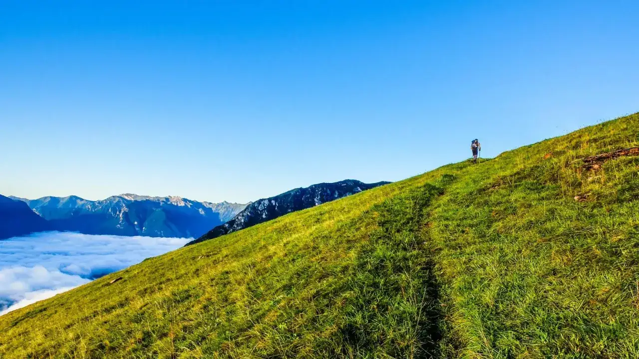 Escursionista solitario sale un prato verdeggiante verso il rifugio Ghedina, con un mare di nuvole sotto e montagne all'orizzonte.