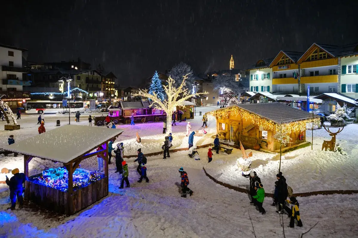 Mercatino di Natale innevato al lago di Andalo, con bancarelle illuminate, alberi addobbati e gente che passeggia.