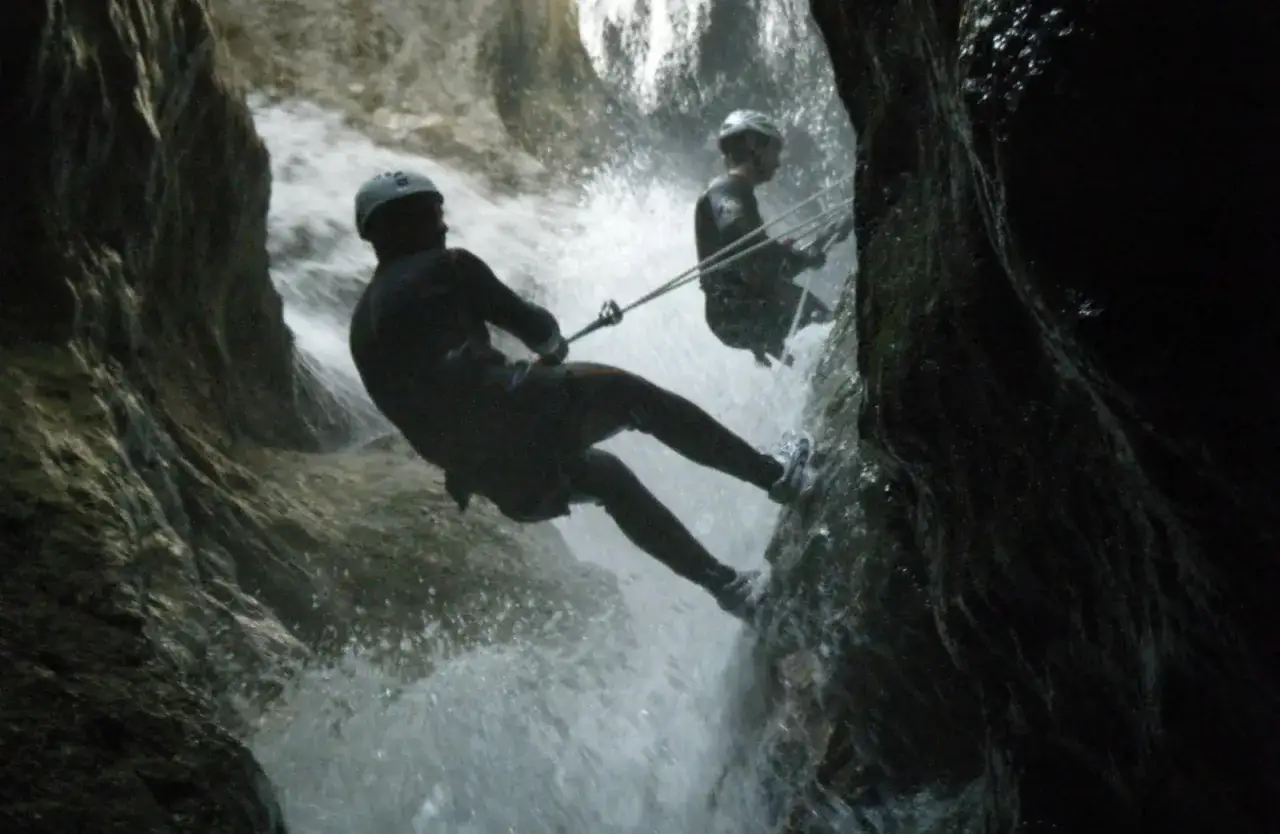 Due persone fanno canyoning in Trentino, calandosi con la corda lungo una cascata tra rocce scure.
