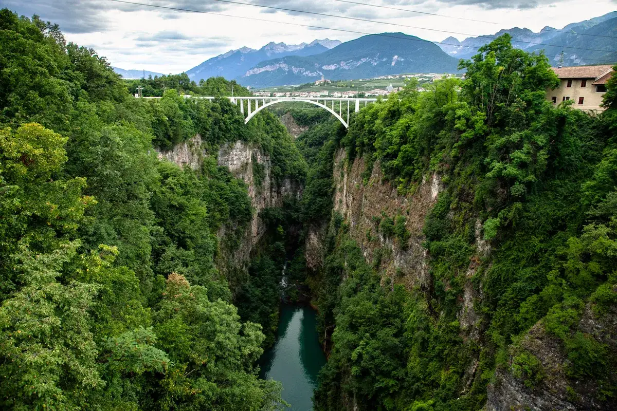 Un ponte bianco attraversa una gola verdeggiante, con il lago della diga di Santa Giustina in basso. Montagne sullo sfondo.